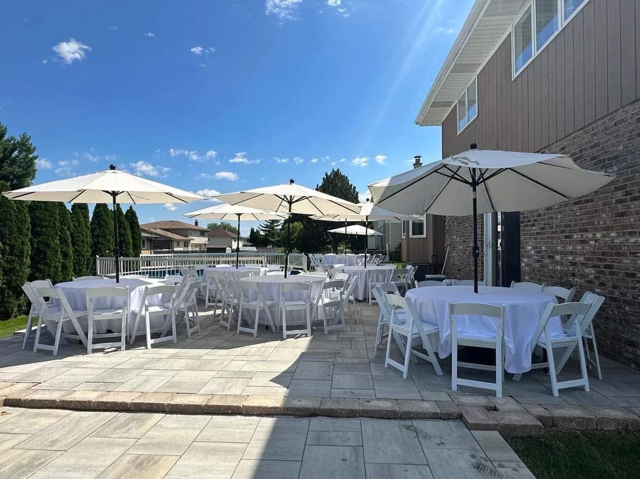 Round tables dressed in white with white folding chairs and cream market umbrellas on a paver patio under a blue sky