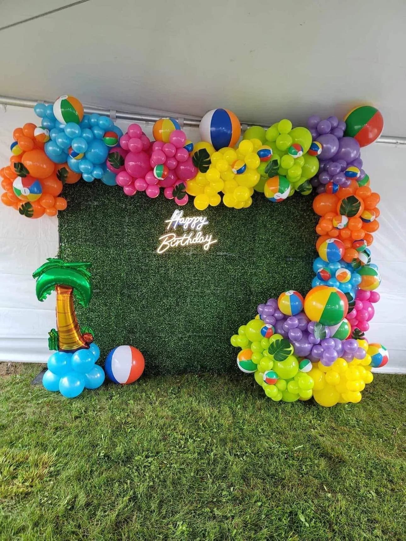 Colorful beach ball balloon garland framing a greenery wall with a Happy Birthday neon sign and a balloon palm tree