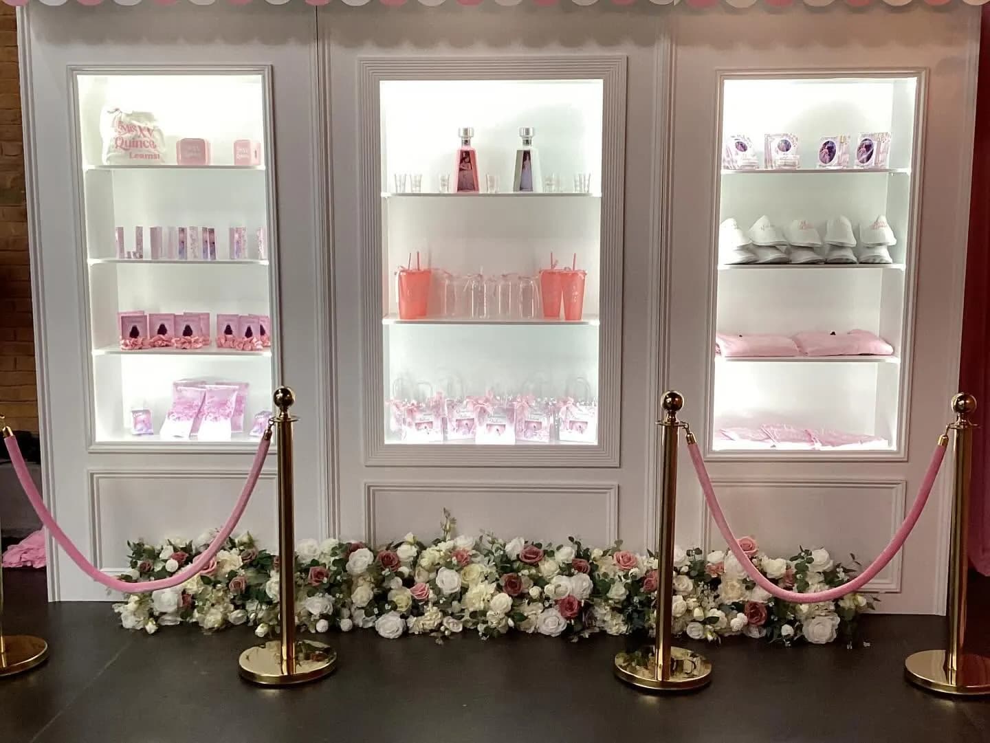 White display wall with three lit niches holding quinceanera favors, a pink striped awning, gold stanchions and a run of white and blush roses