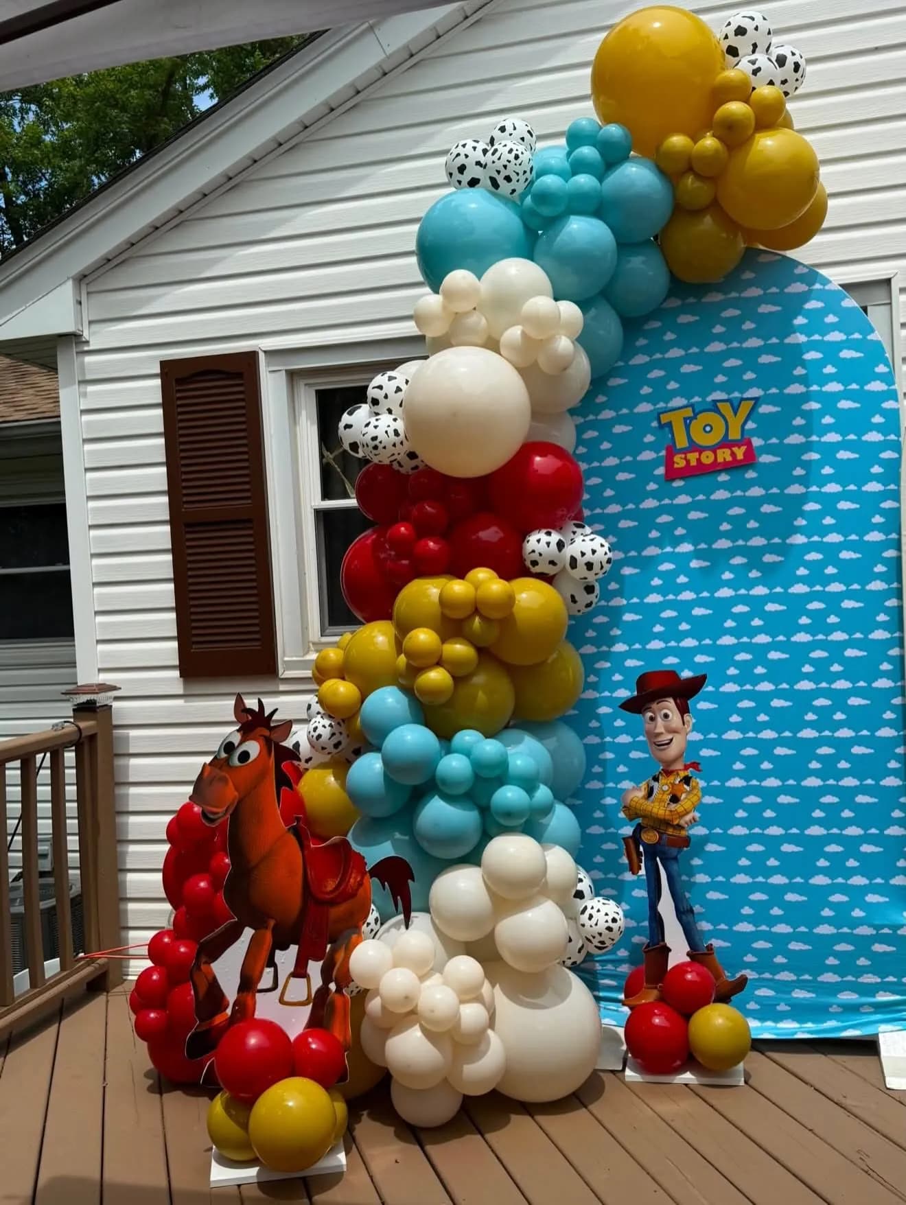 Balloon arch in turquoise, mustard, red and cow print with a cloud backdrop and character cutouts on a wooden deck