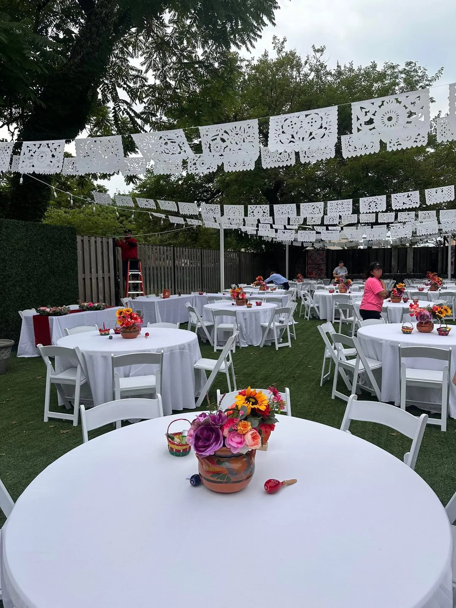 Backyard set with rows of white papel picado overhead, round white tables, folding chairs and flower centerpieces in painted clay pots