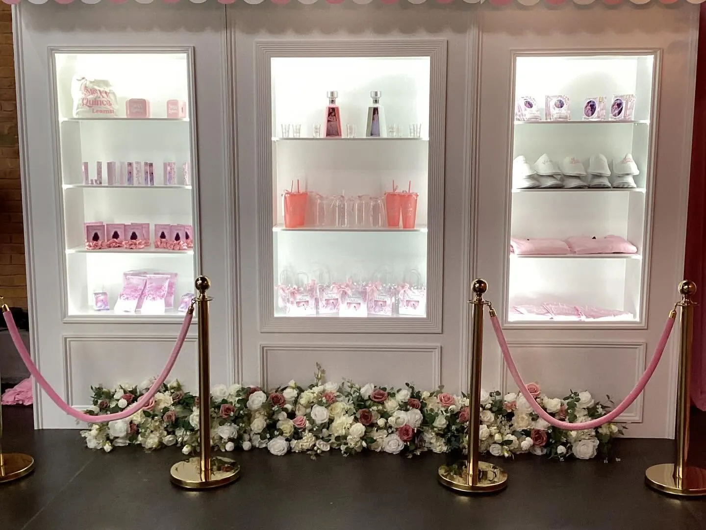 White display wall with three lit niches holding quinceanera favors, a pink striped awning, gold stanchions and a run of white and blush roses